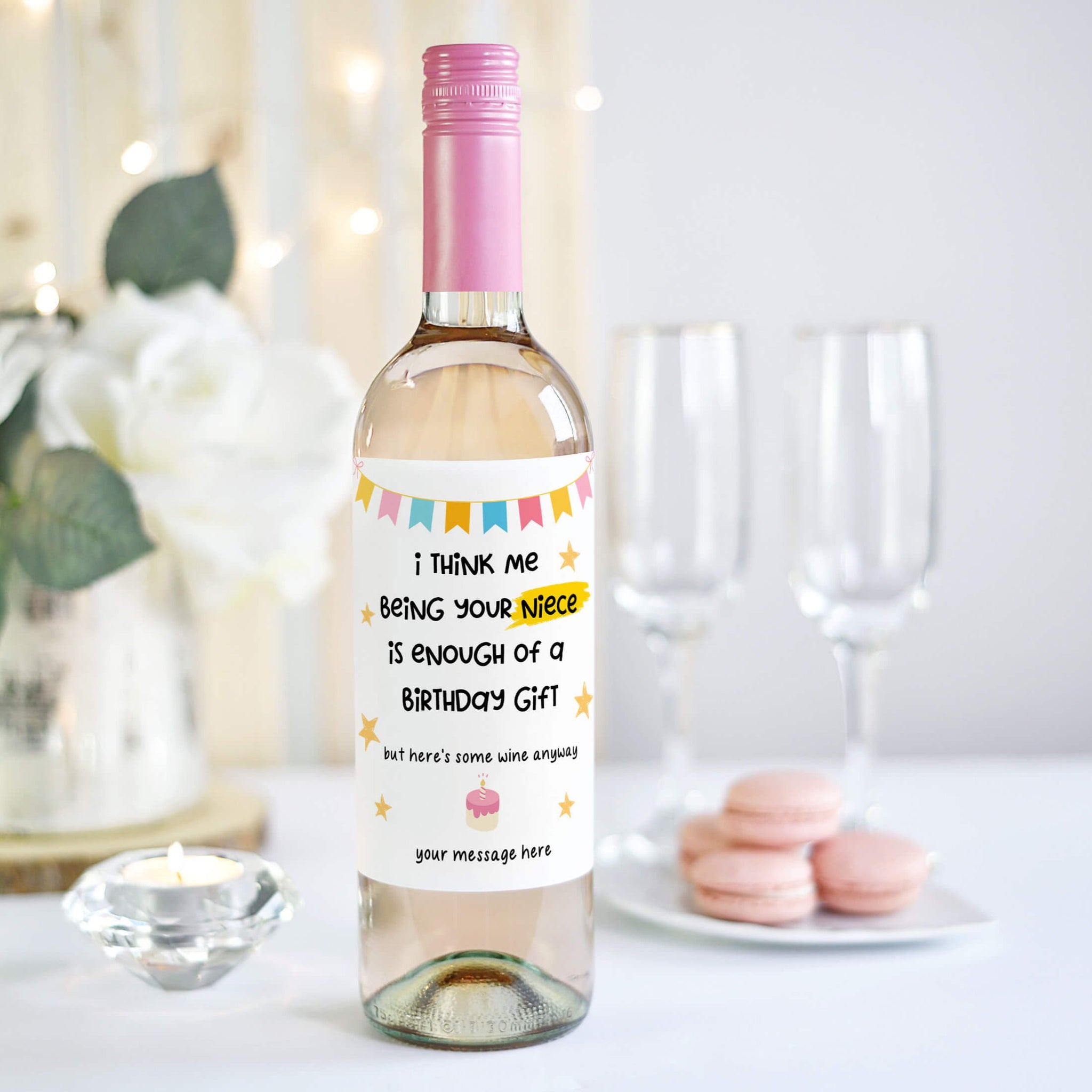 Humorous birthday wine label on a bottle saying 'I think me being your niece is enough of a birthday gift,' styled with pink macarons, white roses, and champagne glasses in the background.